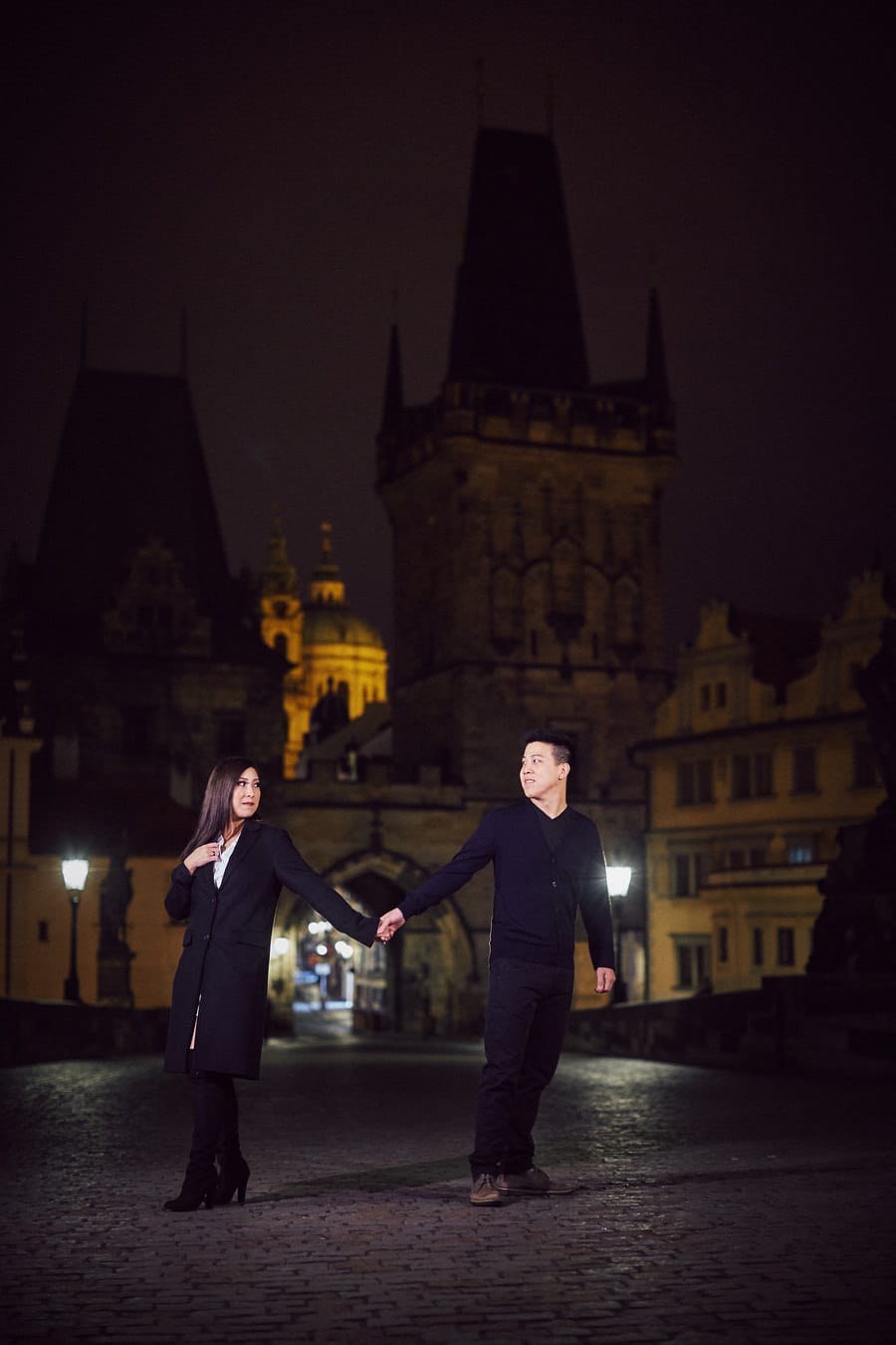 Prague Charles Bridge at night, young couple wearing black, holding hands, gas lamps